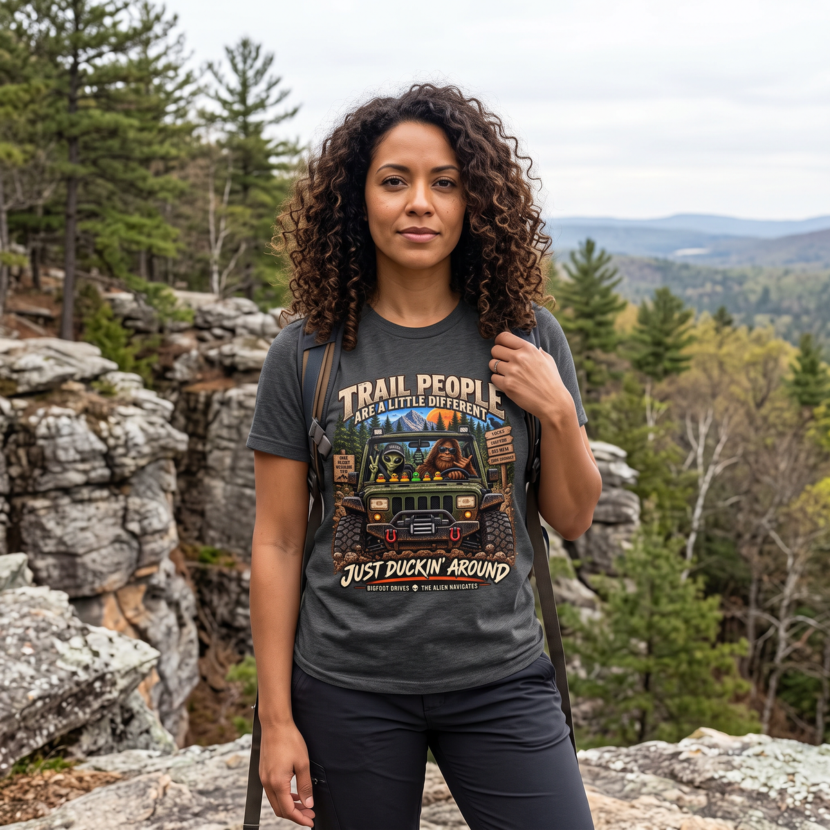 Trail People Dark Heather graphic T-shirt modeled by a curly-haired woman near an outdoor overlook