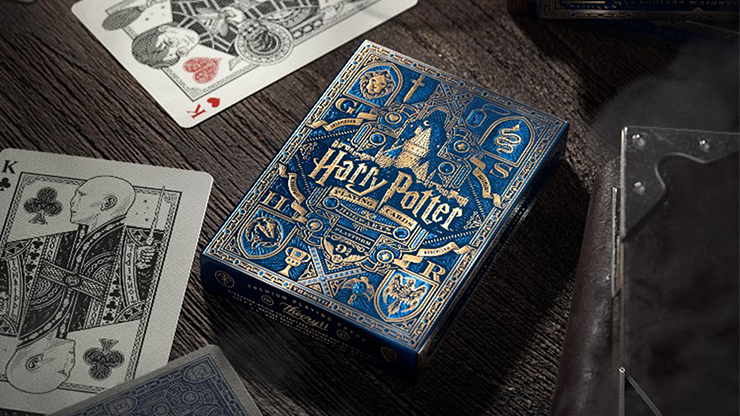 Blue Harry Potter playing card deck by theory11 resting on a wooden table with partially visible cards from the Wizarding World deck.
