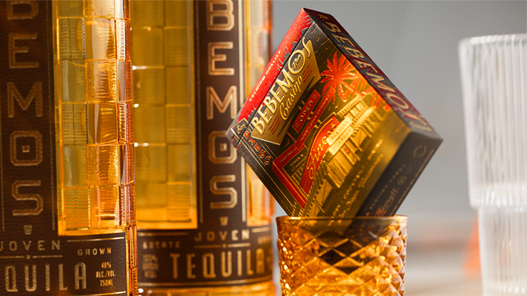Bebemos Casino playing cards by theory11 retro deck tuck case balanced on a crystal glass beside tequila bottles in warm golden bar lighting