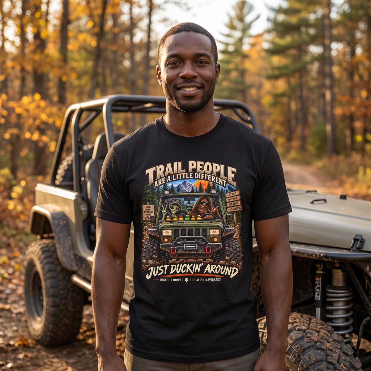 Trail People black graphic T-shirt worn by a model on a wooded Michigan trail, featuring the Bigfoot and alien off-road design