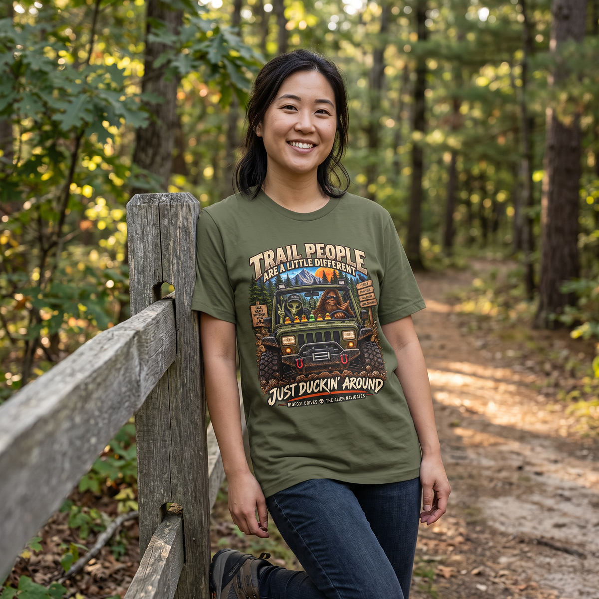 Trail People Military Green graphic T-shirt modeled by a woman near a wooded trail, IMP-POSSIBLE